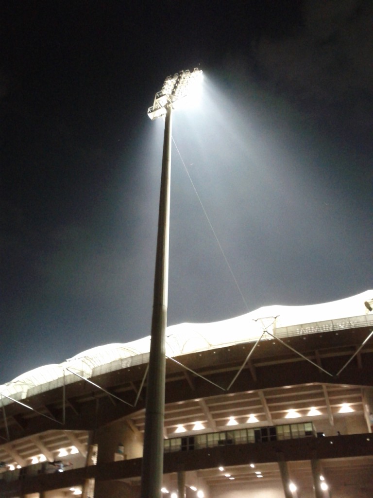 A modern football stadium at night illuminated by powerful floodlights, with digital lines and dots superimposed over the pitch, illustrating player movement tracking and data anal