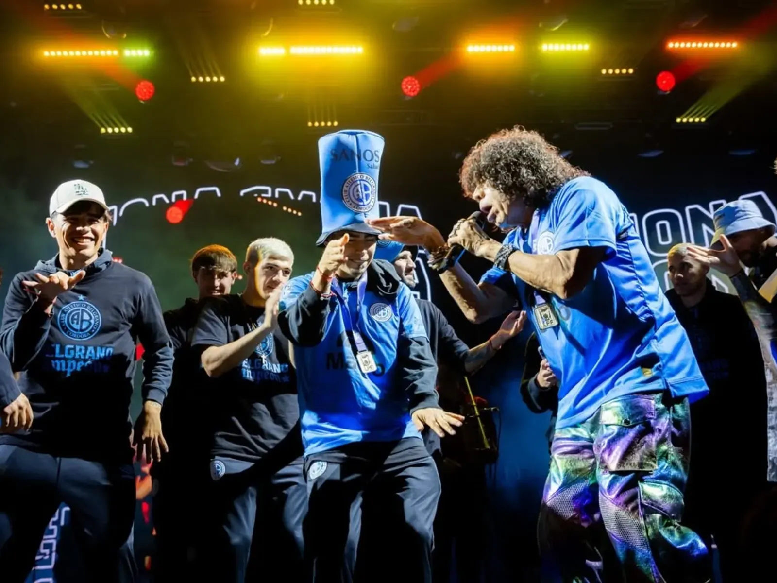 Jugadores de Belgrano celebran en el escenario junto a La Mona Jiménez, portando gorros, banderas y la camiseta del club.