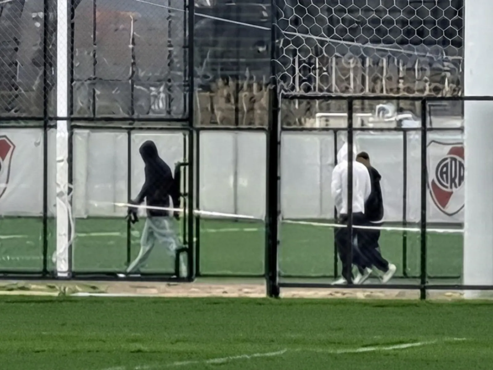 Siete jugadores de River Plate, entre ellos Germán Pezzella, caminan con ropa de entrenamiento hacia una cancha de césped natural en el predio de Cantilo, con containers de obra
