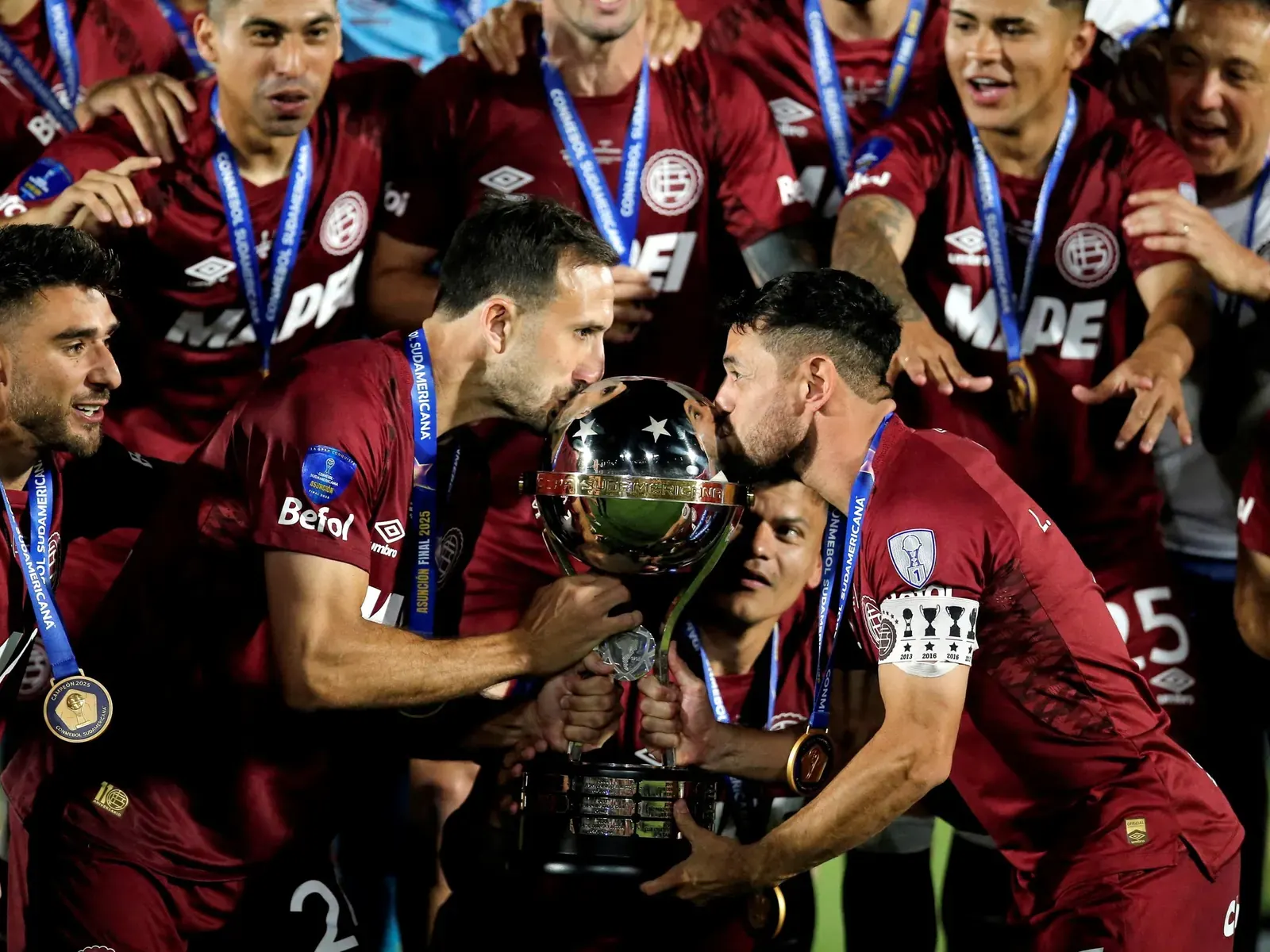 Equipo de Lanús celebrando un gol en un estadio, con hinchas de fondo.